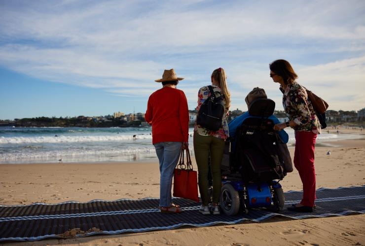 新南威尔士州，悉尼，邦迪海滩，澳大利亚风尚旅行社，一位坐轮椅的男子与其家人一起坐在海滩通道垫上 © 新南威尔士州旅游局版权所有