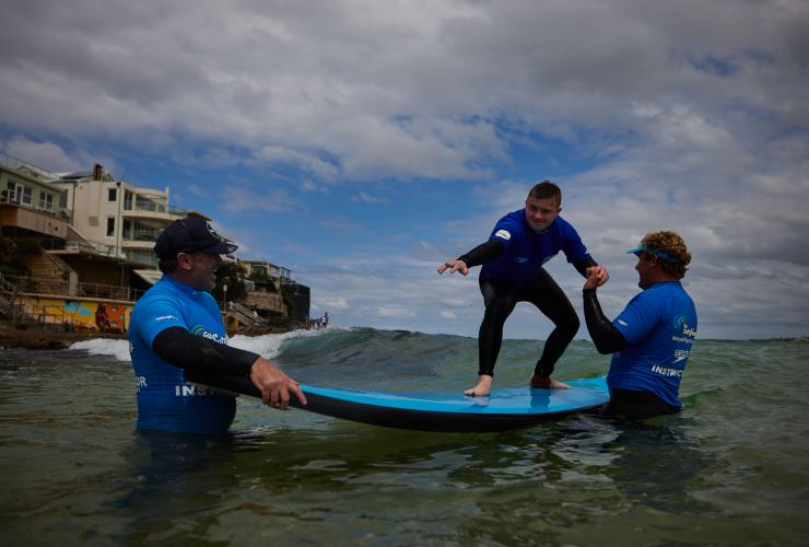 新南威尔士州，悉尼，邦迪海滩，一名患有神经分化症的男子在“海滩冲浪之旅”工作人员的帮助下站在冲浪板上 © 澳大利亚旅游局版权所有