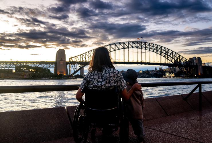 新南威尔士州，悉尼，环形码头，日落时分，一位坐轮椅的男子与其儿子一起眺望悉尼海港大桥 © 澳大利亚旅游局版权所有