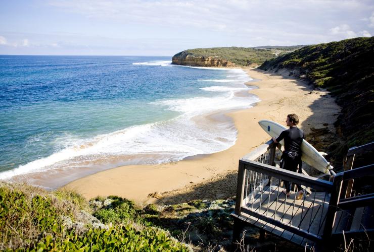 维多利亚州，一名腋下夹着冲浪板的冲浪者站在大洋路一侧的金色海滩上 © 澳大利亚旅游局版权所有