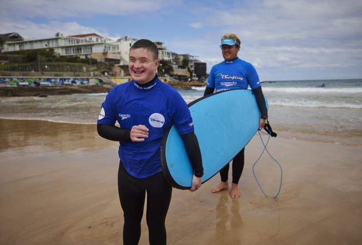 新南威尔士州，悉尼，邦迪海滩上，在教练的指导下，一位患有智力缺陷的年轻男子拿着冲浪板笑逐颜开 © 澳大利亚旅游局版权所有