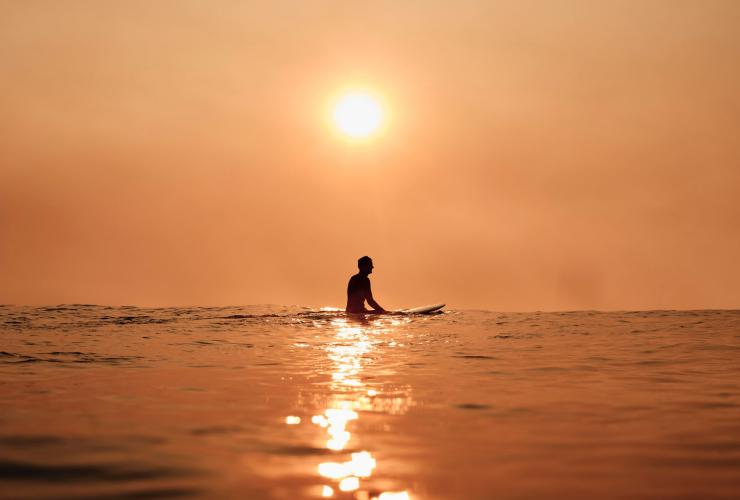 新南威尔士州，悉尼，邦迪海滩上，在橙色晚霞的映照下，可以看到一名冲浪者坐在冲浪板上的身影 © 澳大利亚旅游局版权所有