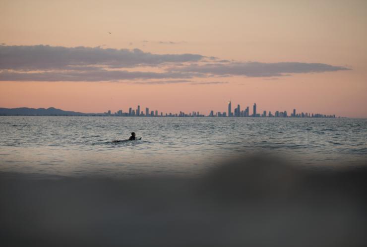 昆士兰州，黄金海岸，一名冲浪者躺在鲷鱼岩海上的冲浪板上，身后是黄金海岸的市区景观 © 昆士兰州旅游及活动推广局版权所有