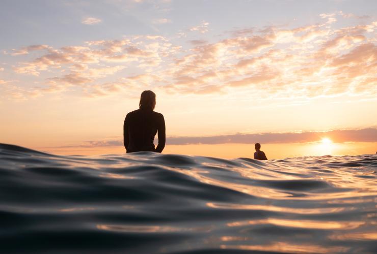 新南威尔士州，拜伦湾上，在朝阳的映照下，两位冲浪者坐在海中的冲浪板上 © 澳大利亚旅游局版权所有