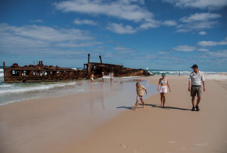 昆士兰州，卡丽岛，两个孩子在翠鸟湾度假村附近的海滩上与导游并肩而行，身后是一群探索岸边沉船残骸的人 © 昆士兰州旅游及活动推广局版权所有