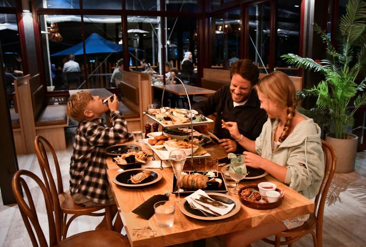 昆士兰州，卡丽岛，翠鸟湾度假村里，一家三口坐在摆满食物的餐桌旁谈笑风生 © 昆士兰州旅游及活动推广局版权所有