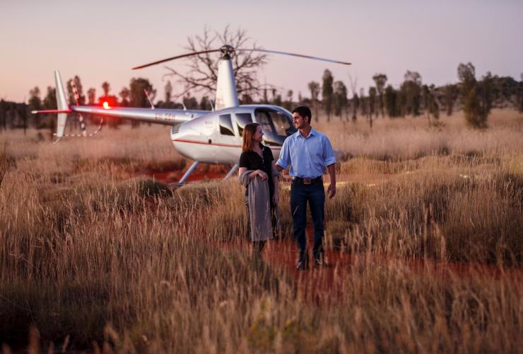 北领地，尤拉拉，东经 131° 酒店，一对情侣在长满草的红土地上漫步，背景是一架直升机 © 澳大利亚旅游局版权所有